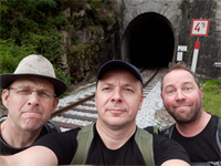 Drei Männer, einer mit Hut und Brille, einer mit einer Kappe und einer mit Bart stehen neben Bahngleisen vor einem Zugtunnel